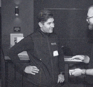 A founder in a dark vest and name tag, hand on his hip, smiling at another member across a bar counter