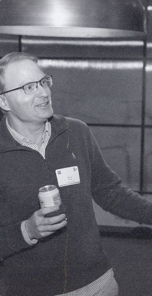 A founder in glasses and a quarter-zip sweater, name tag on and a can in hand, smiling as he talks at a club evening