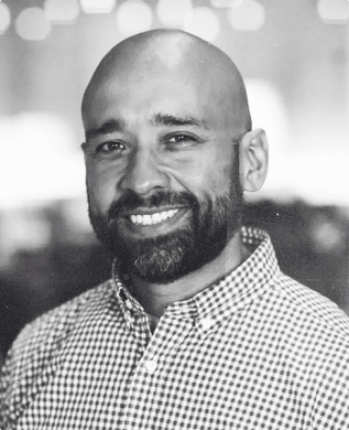 David Cancel in a checked shirt, smiling, against blurred lights