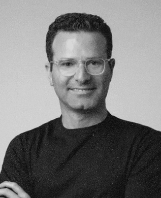Eran Ben-Shushan in round glasses and a black T-shirt, arms crossed, against a grey backdrop