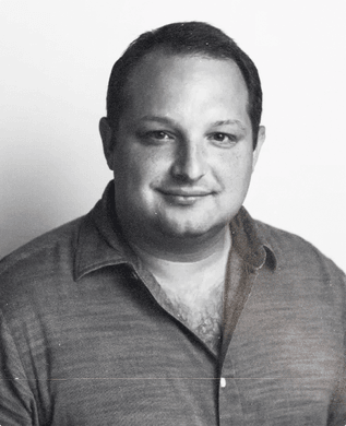 David Politis in an open-collared shirt, smiling, against a pale wall