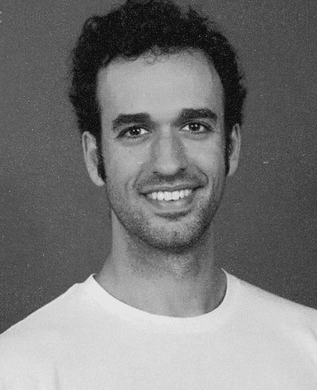 Miguel Fernandez with dark curly hair, smiling, in a white T-shirt against a grey backdrop