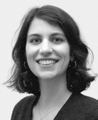 A smiling woman with shoulder-length dark wavy hair and hoop earrings, in a dark cardigan against a pale wall