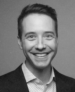 Zachary Smith, smiling, in a dark blazer over a patterned shirt against a grey backdrop