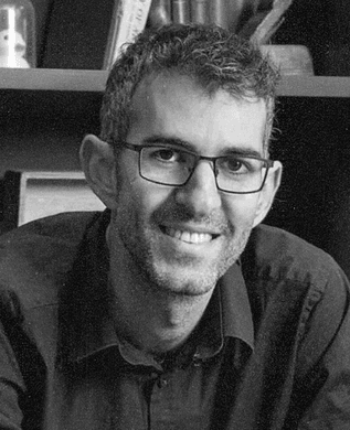 Nitzan Shapira in glasses and a dark shirt, smiling, in front of a bookshelf