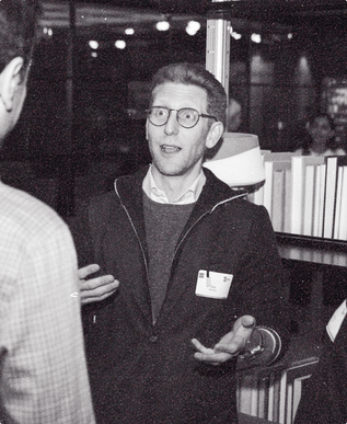 A founder in round glasses and a name tag explains a point, palms open, beside a bookshelf at the December event